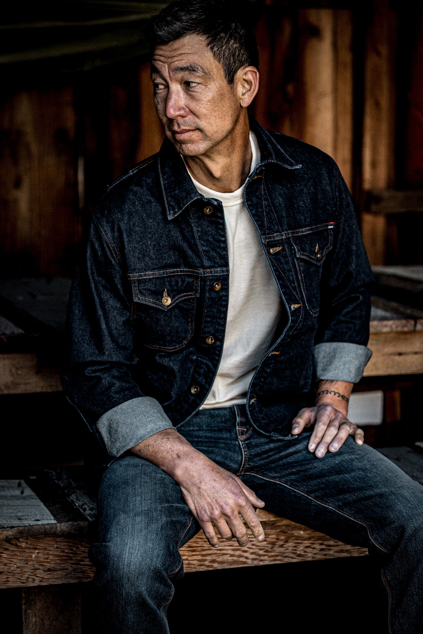 Man in dark broken twill denim jacket with cuffed sleeves and brass buttons, seated on a wooden bench.