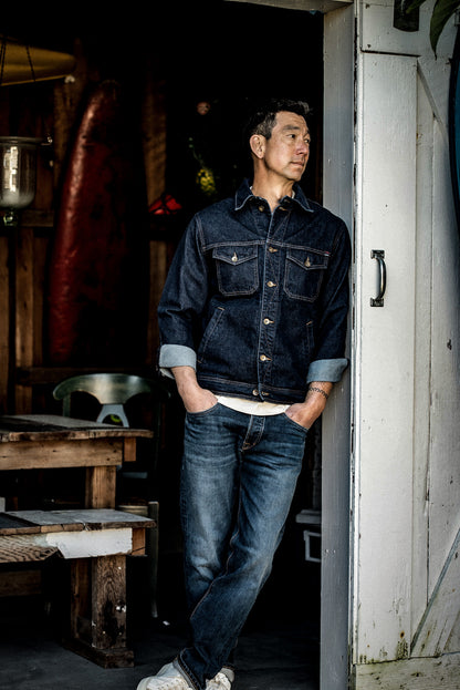 Man wearing the Ocean Artcher denim jacket, cuffs rolled, leaning against a weathered doorway.