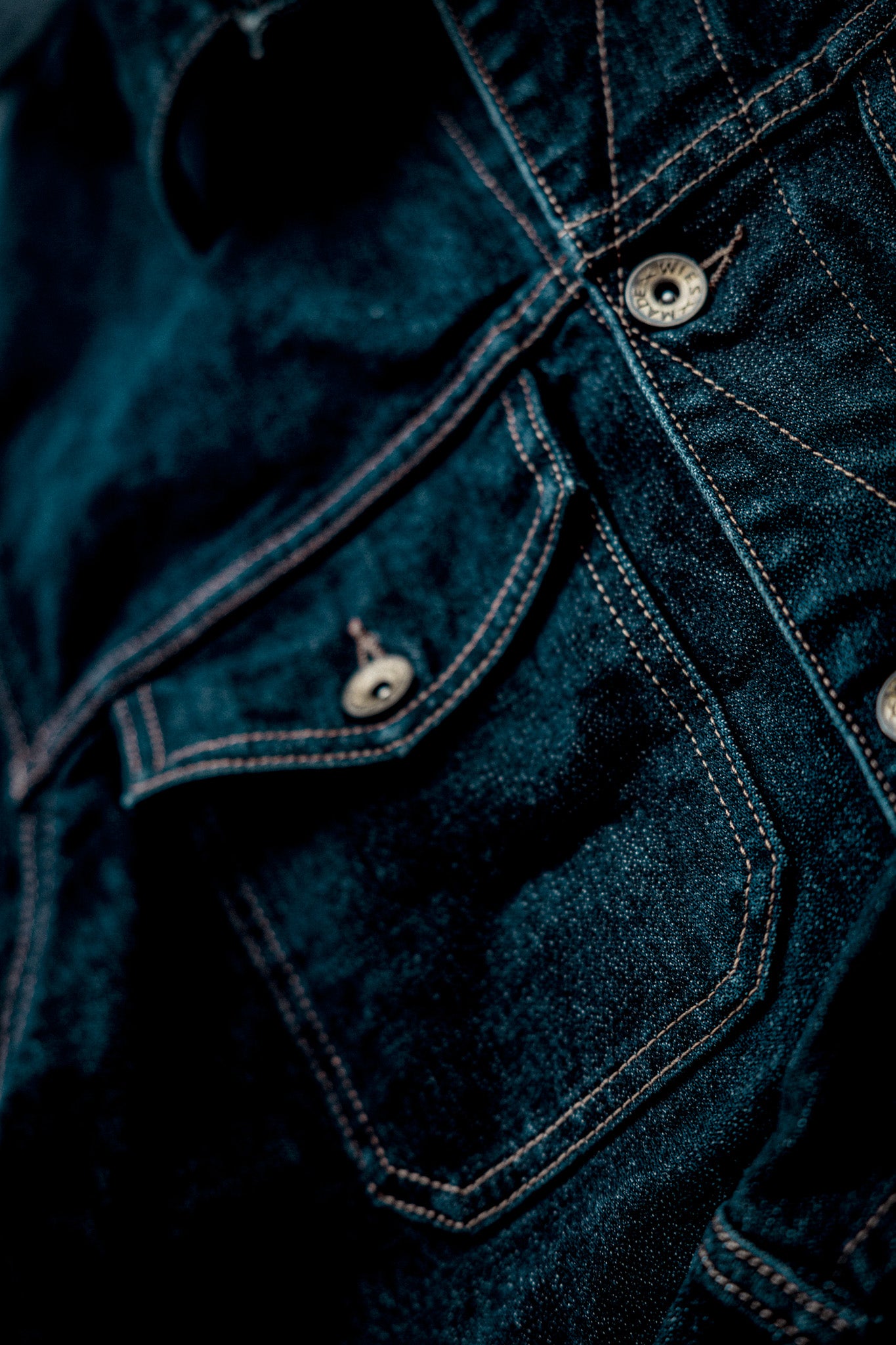Close-up of broken twill denim jacket showing chest pocket, contrast stitching, and engraved brass buttons.