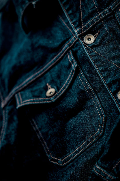 Close-up of broken twill denim jacket showing chest pocket, contrast stitching, and engraved brass buttons.