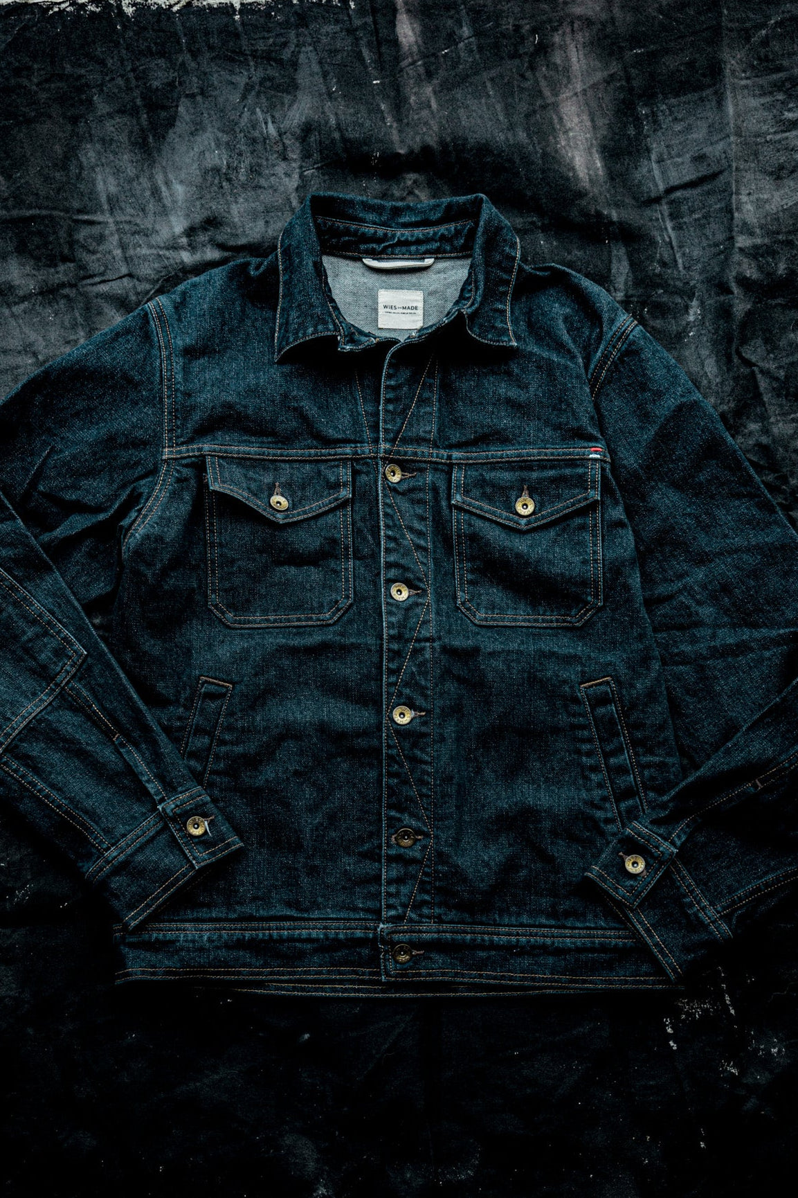 Men's broken twill denim jacket in dark ocean blue, laid flat with brass buttons and chest pockets.