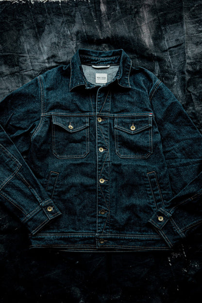 Men's broken twill denim jacket in dark ocean blue, laid flat with brass buttons and chest pockets.