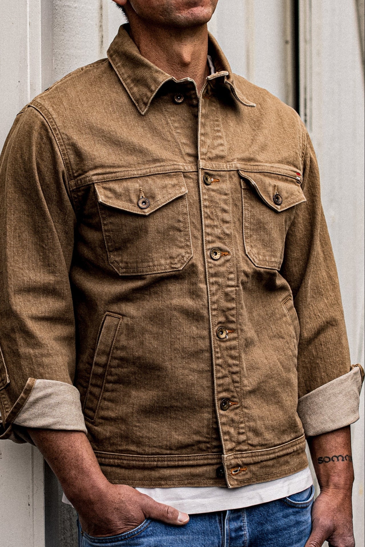 Close-up of the Artcher wheat broken twill denim jacket worn with rolled cuffs, chest flap pockets, and brass buttons.