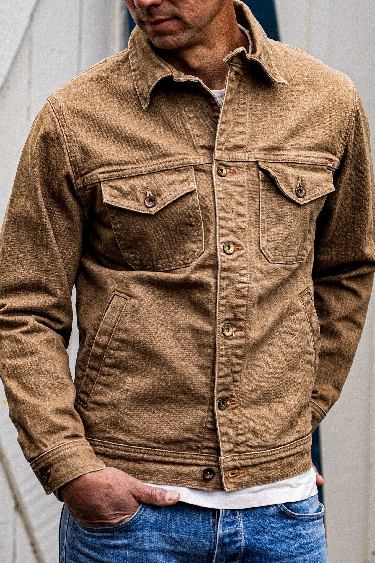 Man wearing a wheat-colored broken twill denim jacket with chest flap pockets and brass buttons.