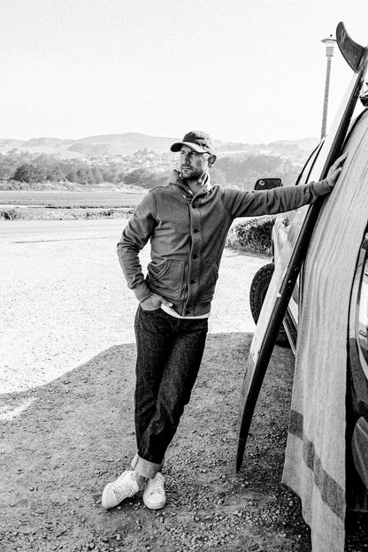 Man wearing button-up Bodega French terry cardigan and cuffed raw denim beside a truck.