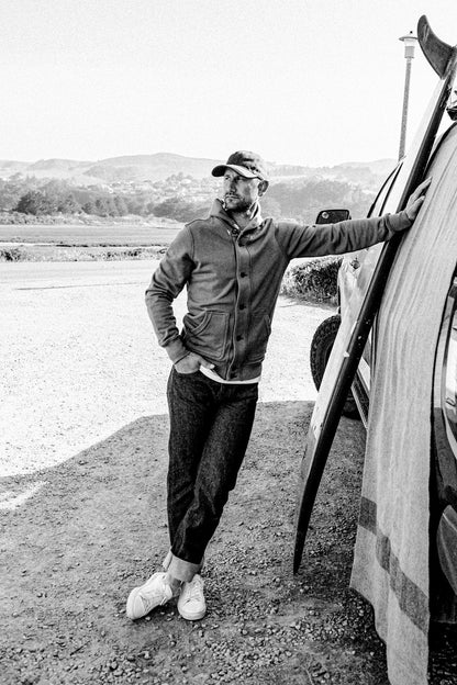 Man in Bodega French terry cardigan and raw denim jeans leaning on a truck with a surfboard near the coast.