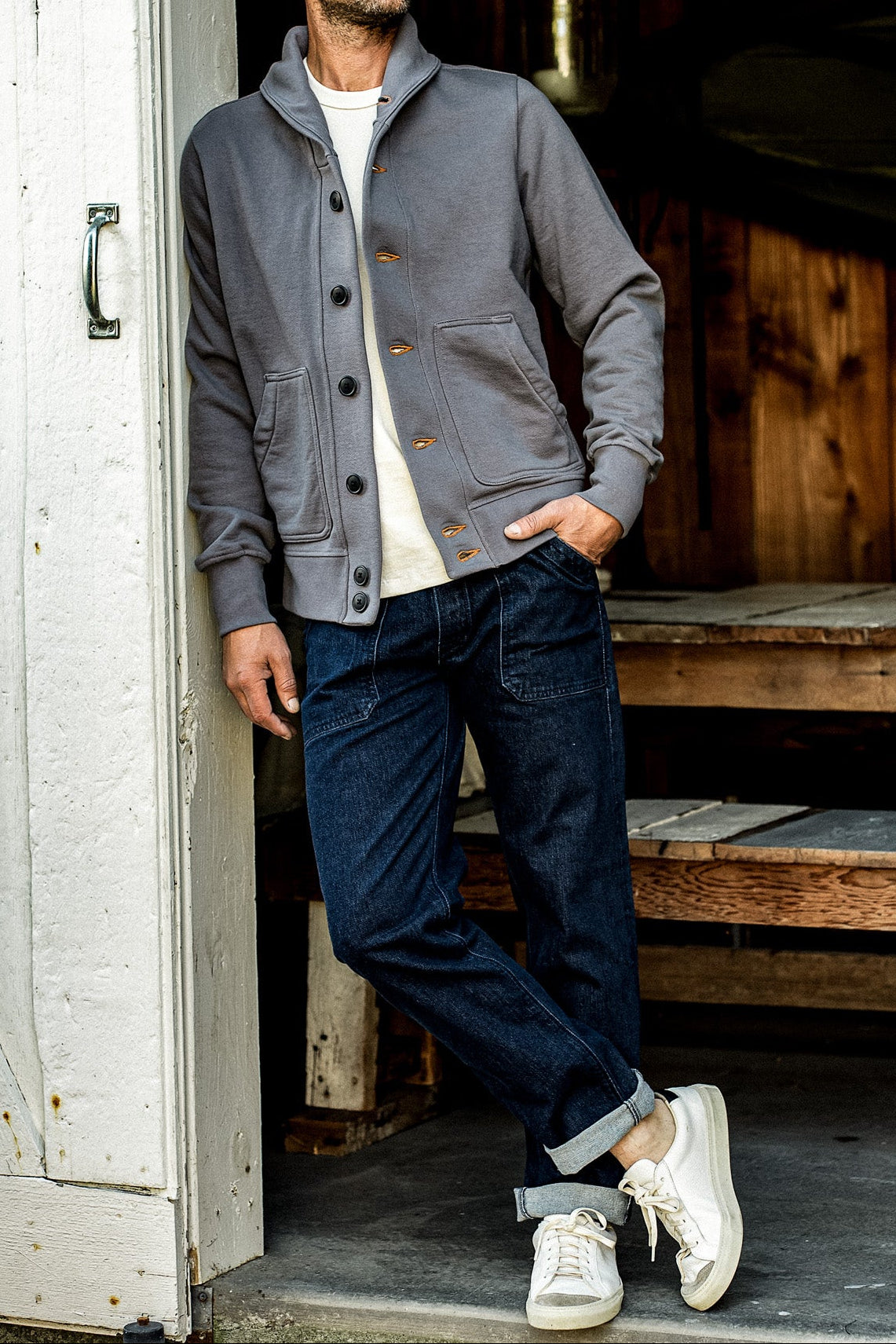 Full body shot of man in the WiesMade Bodega grey cardigan open with dark rolled-cuff jeans and white sneakers.