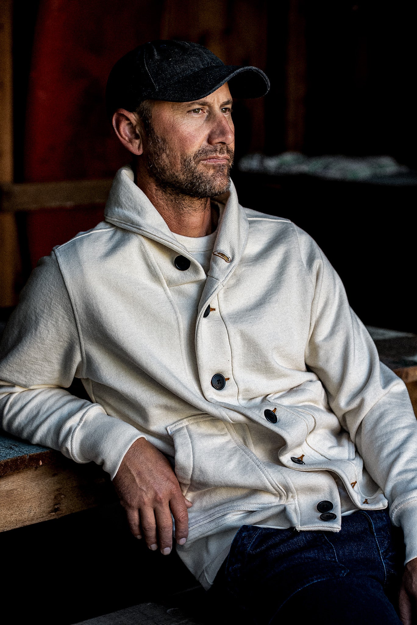 Man in natural French terry Bodega cardigan with contrast buttons, seated in a rustic barn.