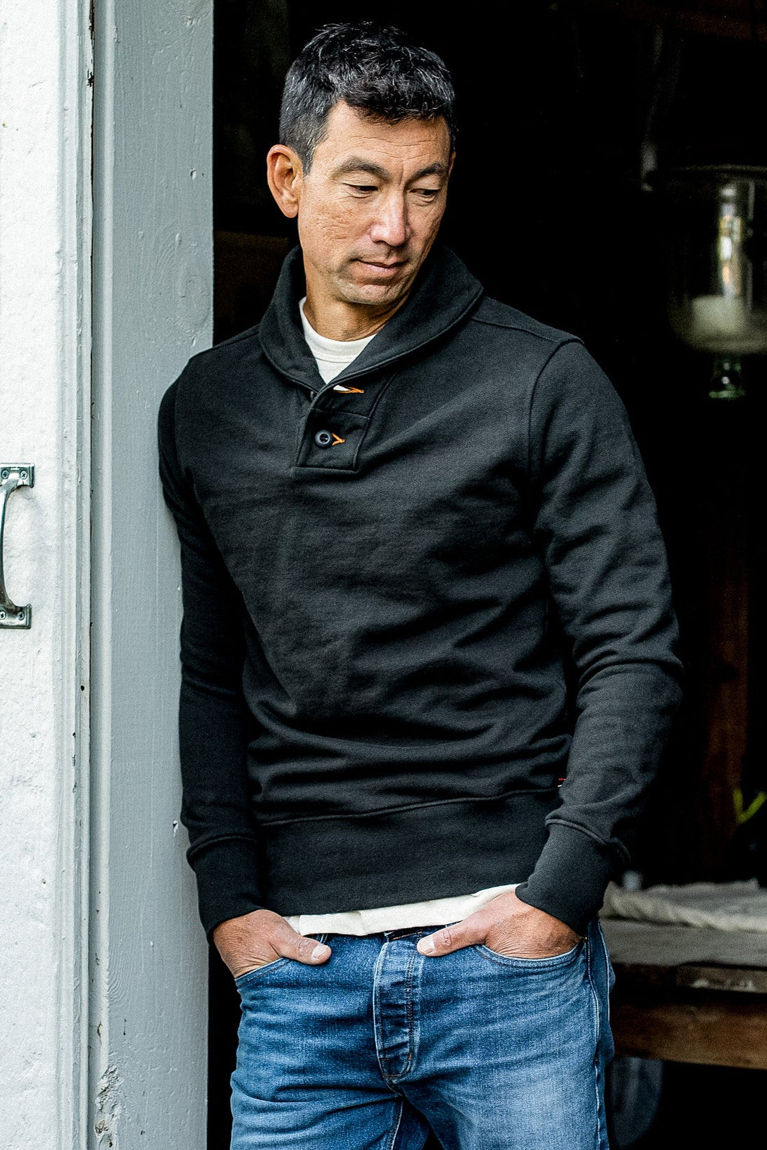 Man in a basalt black shawl collar French terry pullover and blue jeans leaning in a doorway.