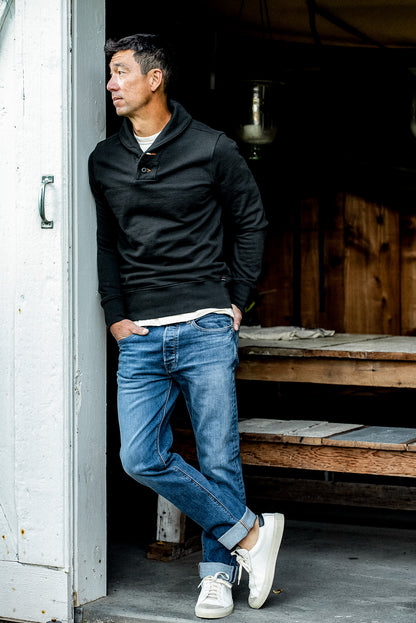 Man in basalt black shawl collar French terry pullover and cuffed blue jeans leaning in a doorway.