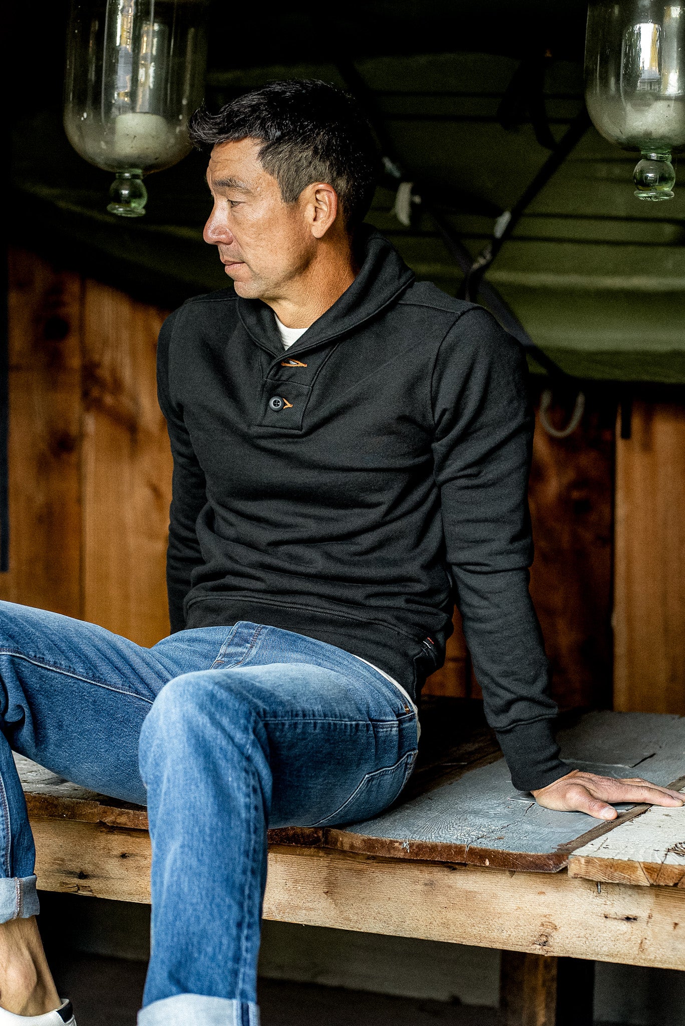 Man in a basalt shawl collar French terry pullover and blue jeans seated on a rustic wooden platform.