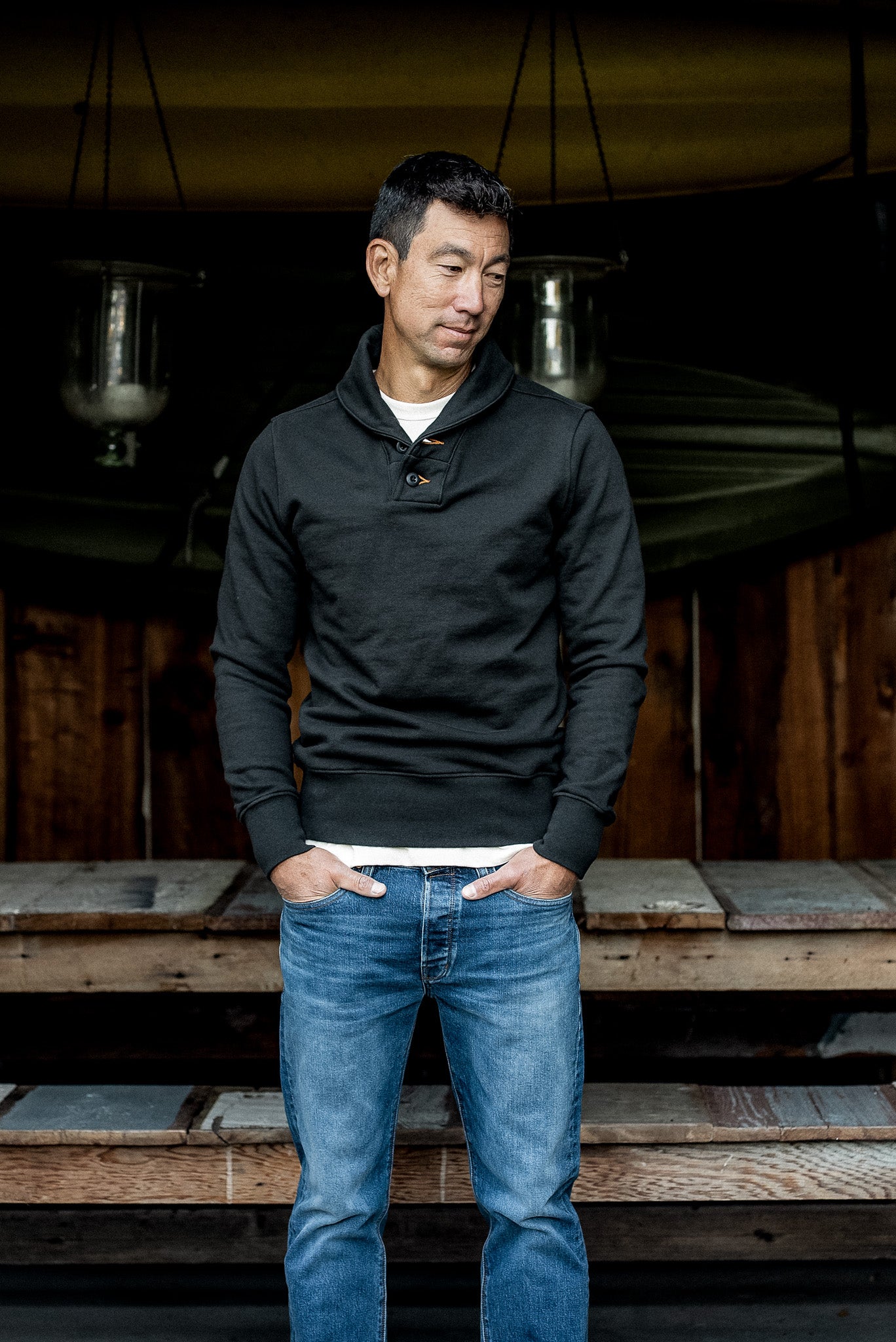 Man wearing a basalt black shawl collar French terry pullover with blue jeans on wooden porch steps.