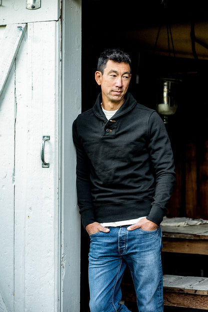 Man in a basalt black shawl collar French terry pullover and blue jeans leaning in a weathered barn doorway.