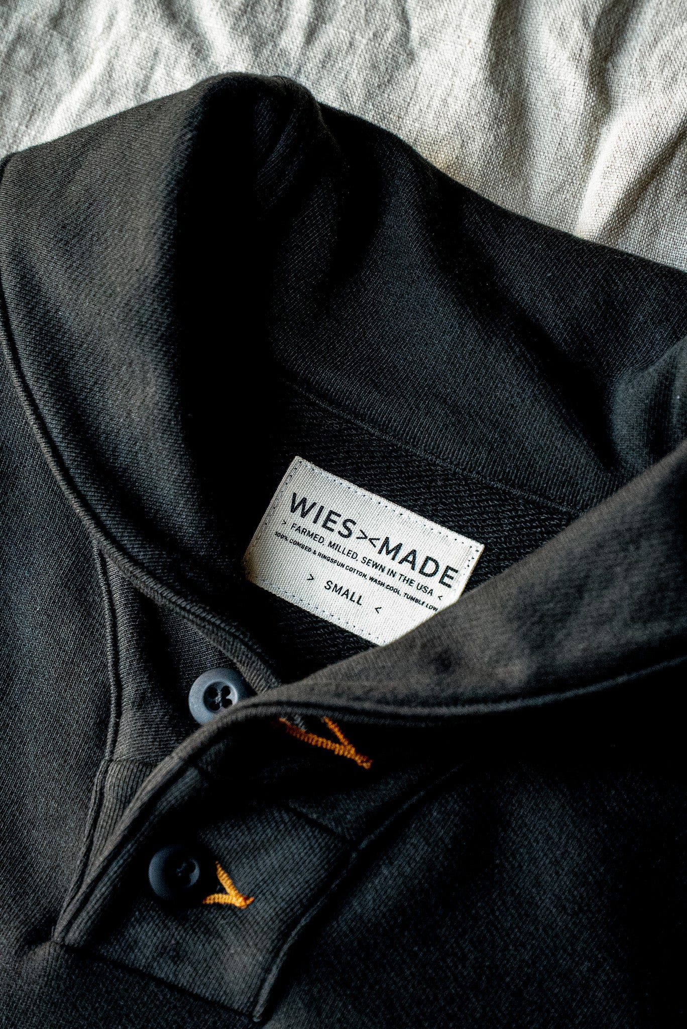 Close-up of basalt French terry shawl collar pullover showing WiesMade label, button placket, and orange contrast stitching.