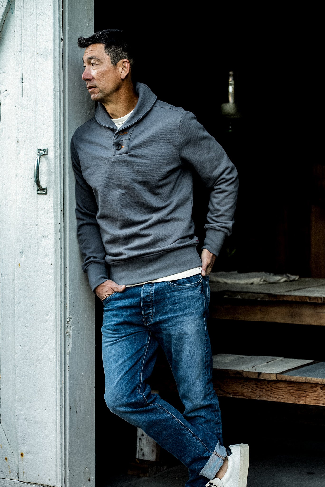 Man wearing the WiesMade Bodega grey pebble shawl-neck pullover with jeans and white sneakers in a doorway.