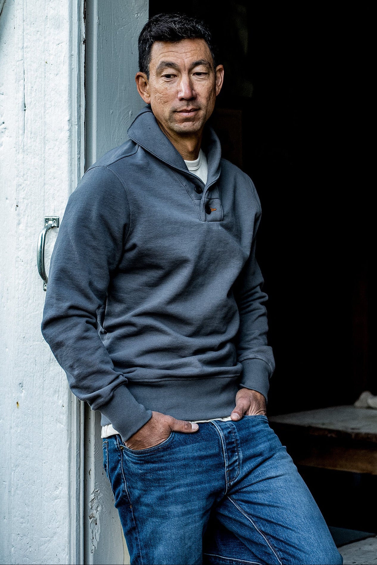 Man in a pebble grey shawl collar pullover leaning against a white doorway with jeans.