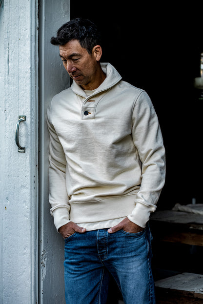 Man in natural cream shawl collar French terry pullover and blue denim jeans leaning in a doorway.