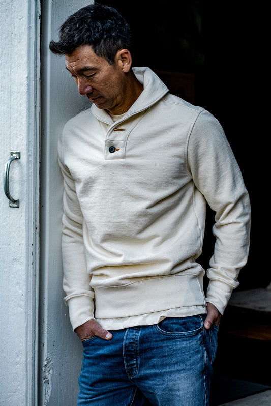Man in a natural cream French terry shawl collar pullover and blue denim jeans, leaning against a weathered doorway.