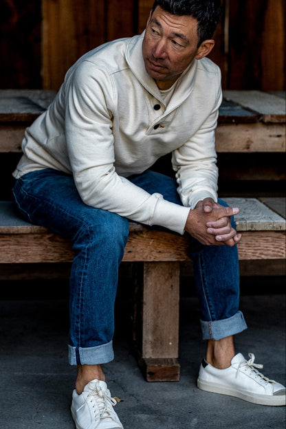 Man in a natural cotton French terry shawl collar pullover sits on a wooden bench with cuffed selvedge denim jeans.