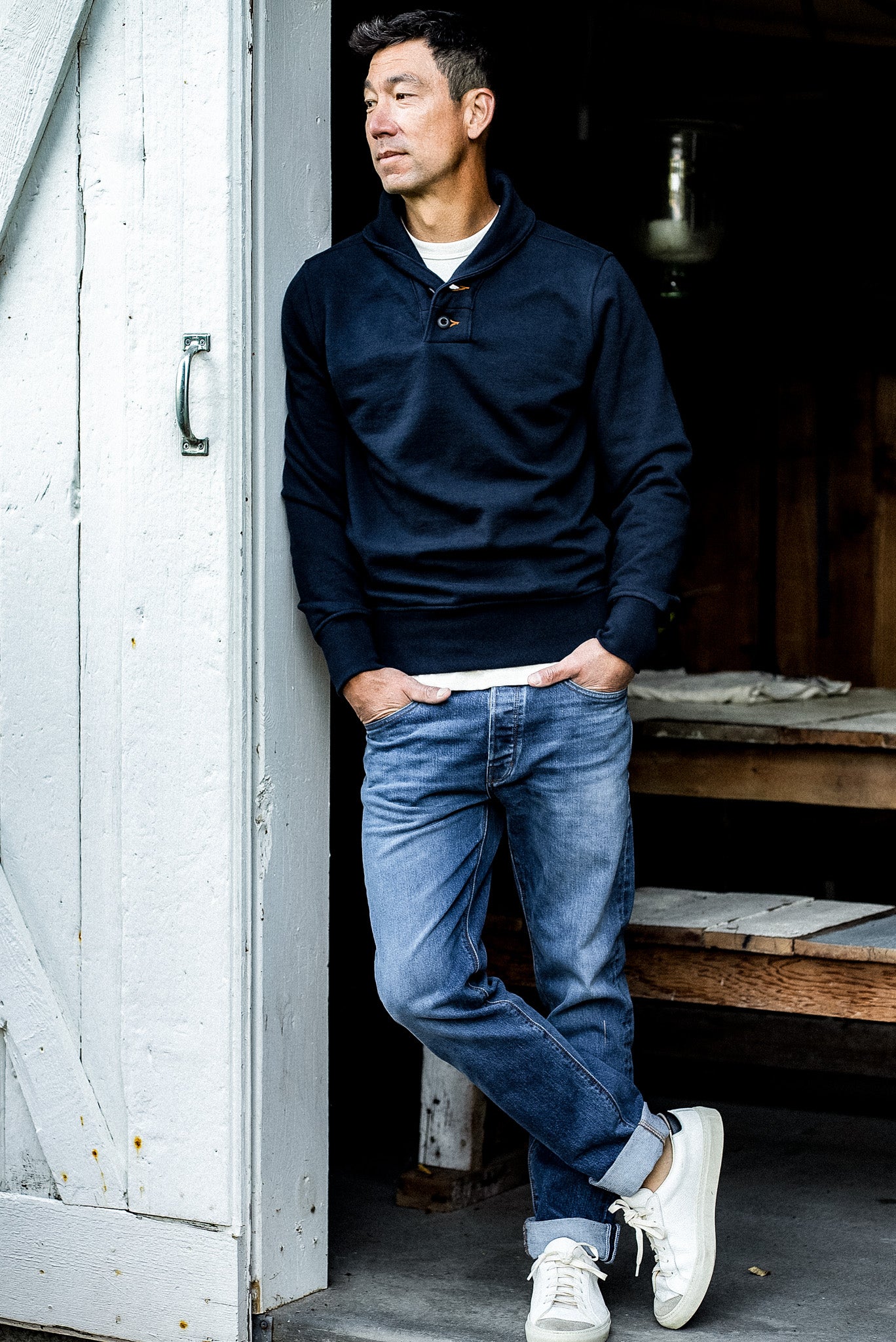 Navy shawl collar French terry pullover worn with jeans, leaning against a white barn door.