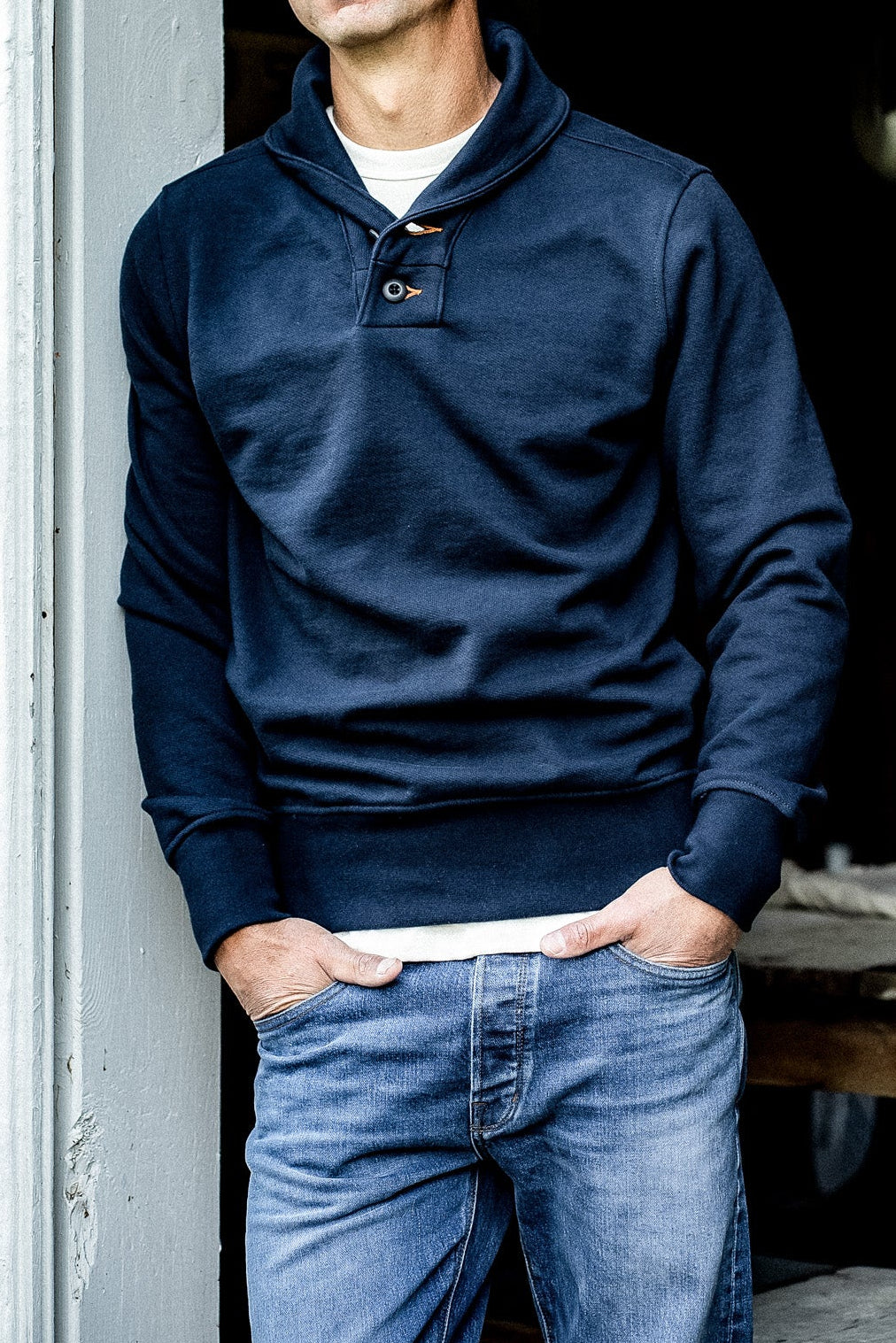 Man in navy shawl collar French terry pullover with button placket, hands in jean pockets.