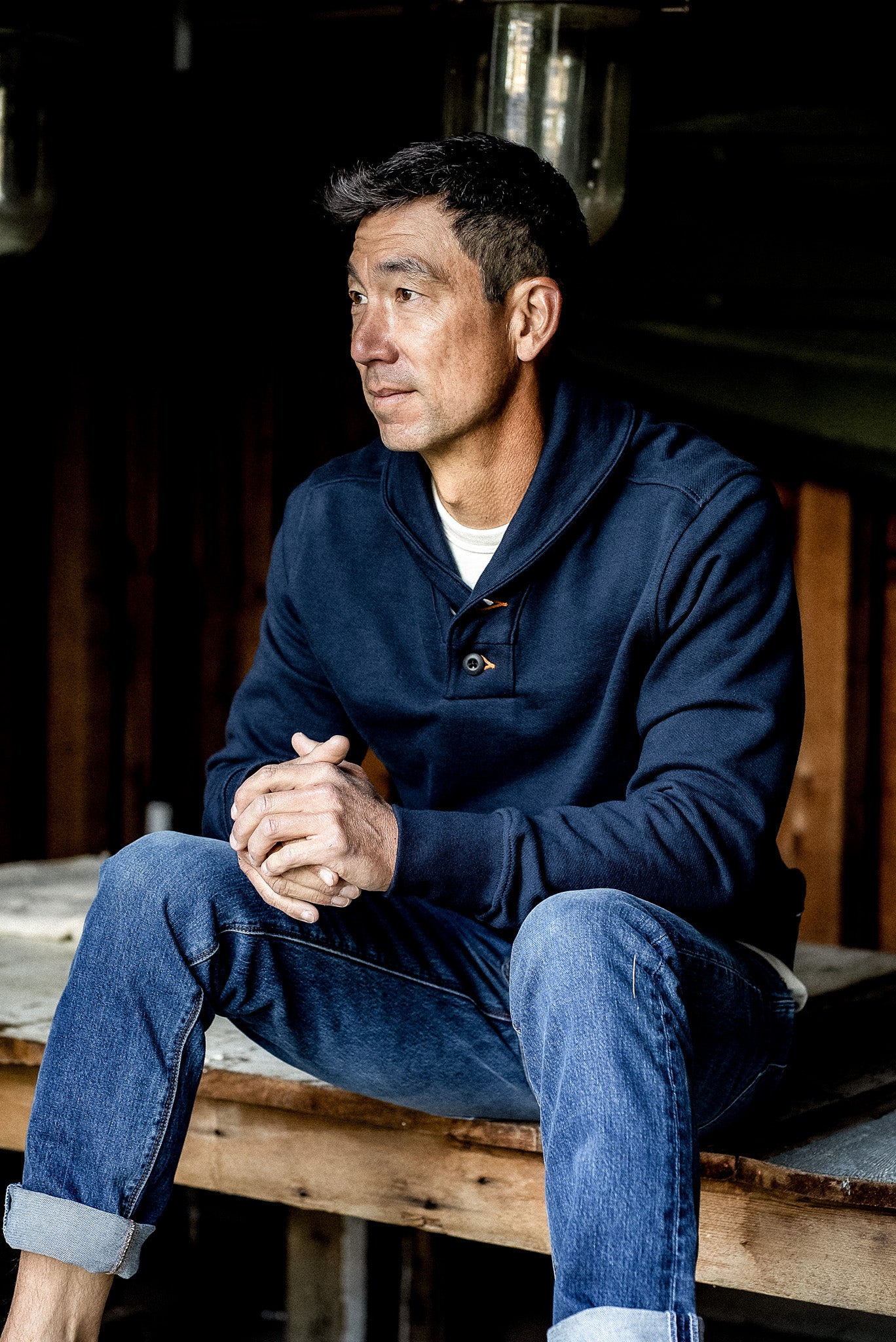 Man wearing navy shawl collar French terry pullover, seated on a wooden bench.