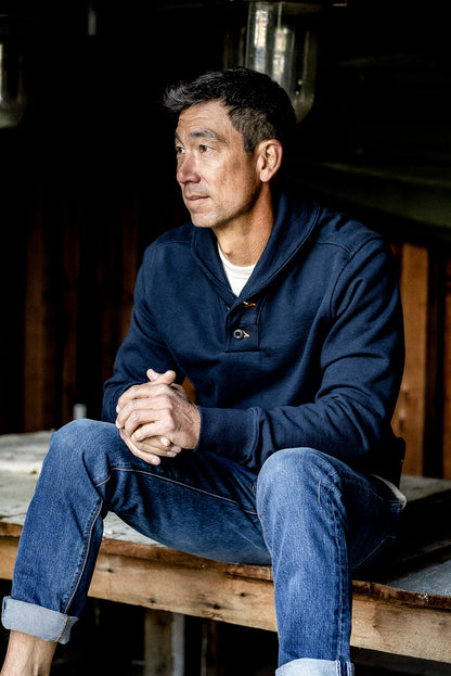 Man wearing navy shawl collar French terry pullover, seated on a wooden bench.
