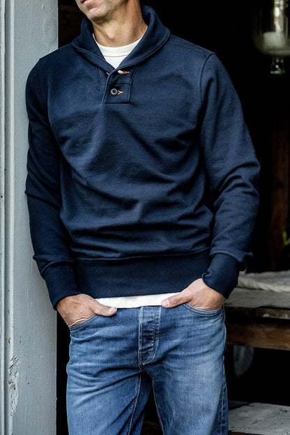 Man wearing the WiesMade Bodega navy shawl-neck pullover with jeans, hands in pockets, leaning against a wall.