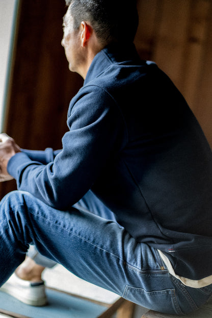 Man sitting on a bench in a WiesMade Bodega navy pullover with jeans, shown from back-side angle.