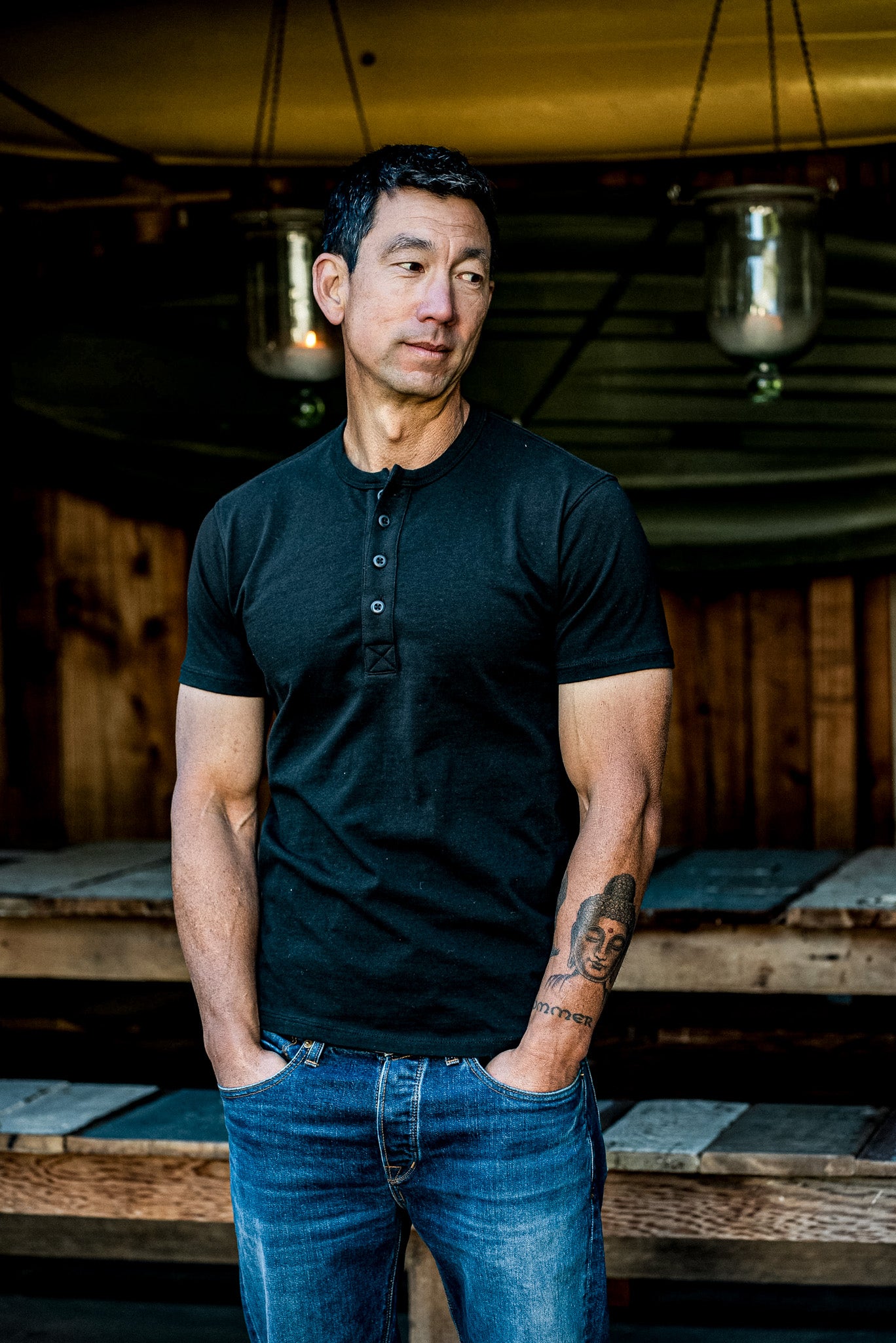 Man wearing the Bolinas four-button Henley in dark Basalt with blue denim jeans, standing on a wooden porch.