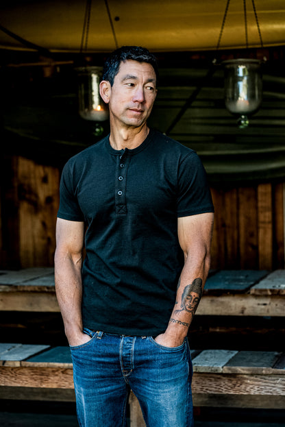 Man wearing the Bolinas four-button Henley in dark Basalt with blue denim jeans, standing on a wooden porch.