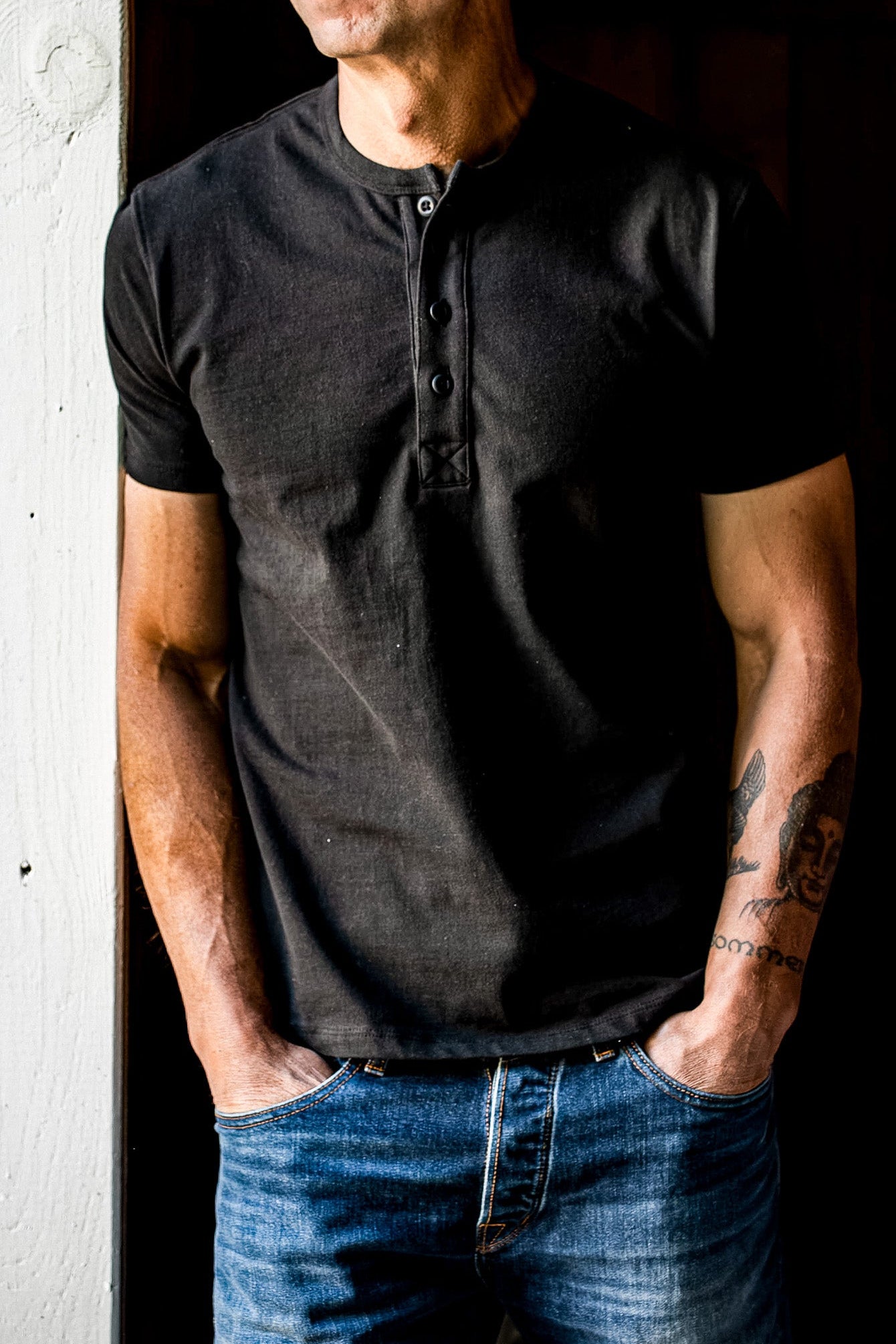 Basalt black four-button Bolinas Henley worn with blue jeans, hands in pockets against a weathered doorway.