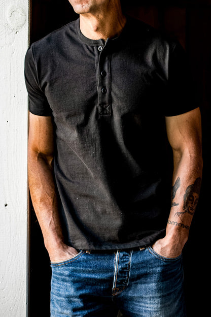 Basalt black four-button Bolinas Henley worn with blue jeans, hands in pockets against a weathered doorway.