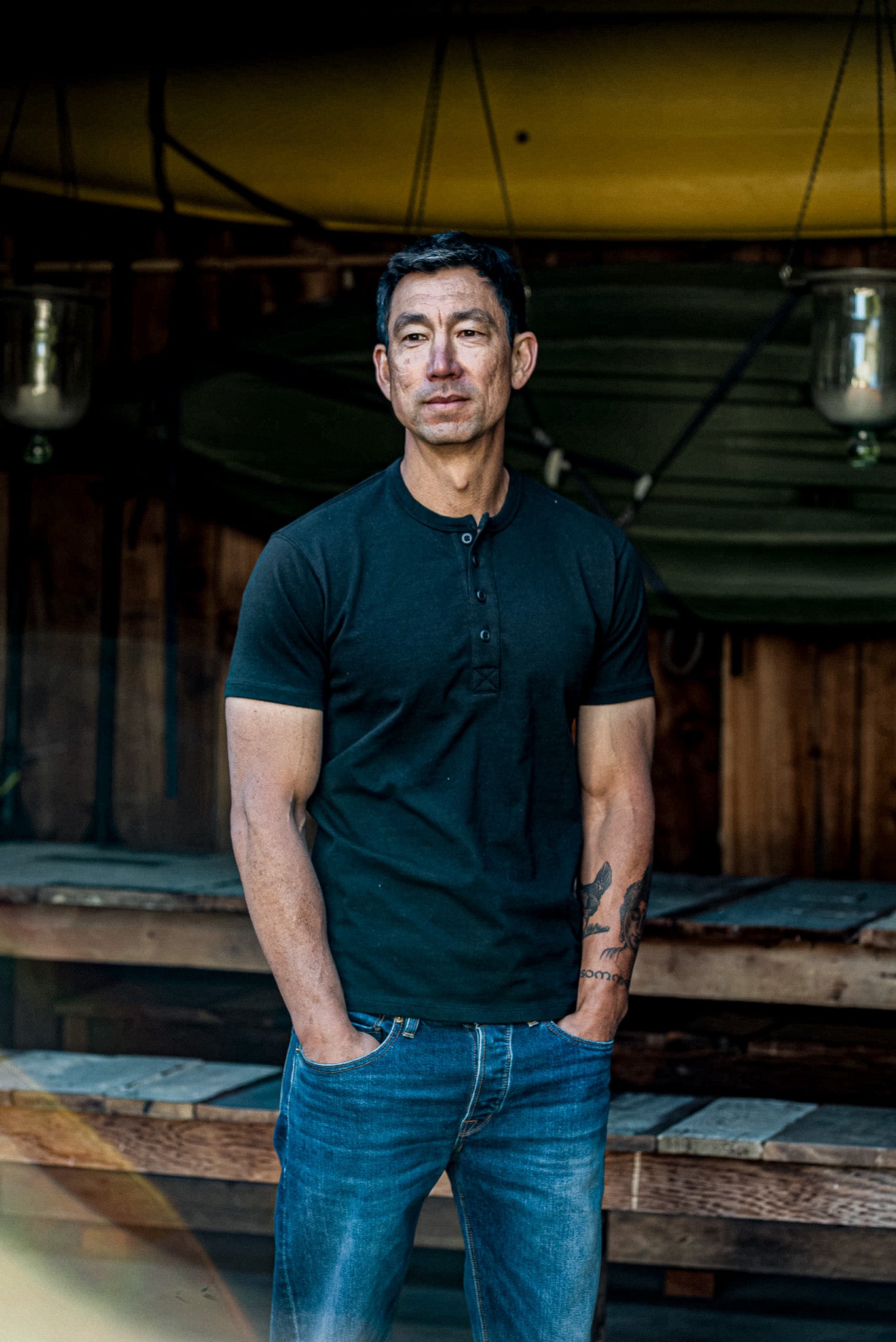 Man in a dark basalt short-sleeve Bolinas henley and blue jeans standing by a wooden boathouse.