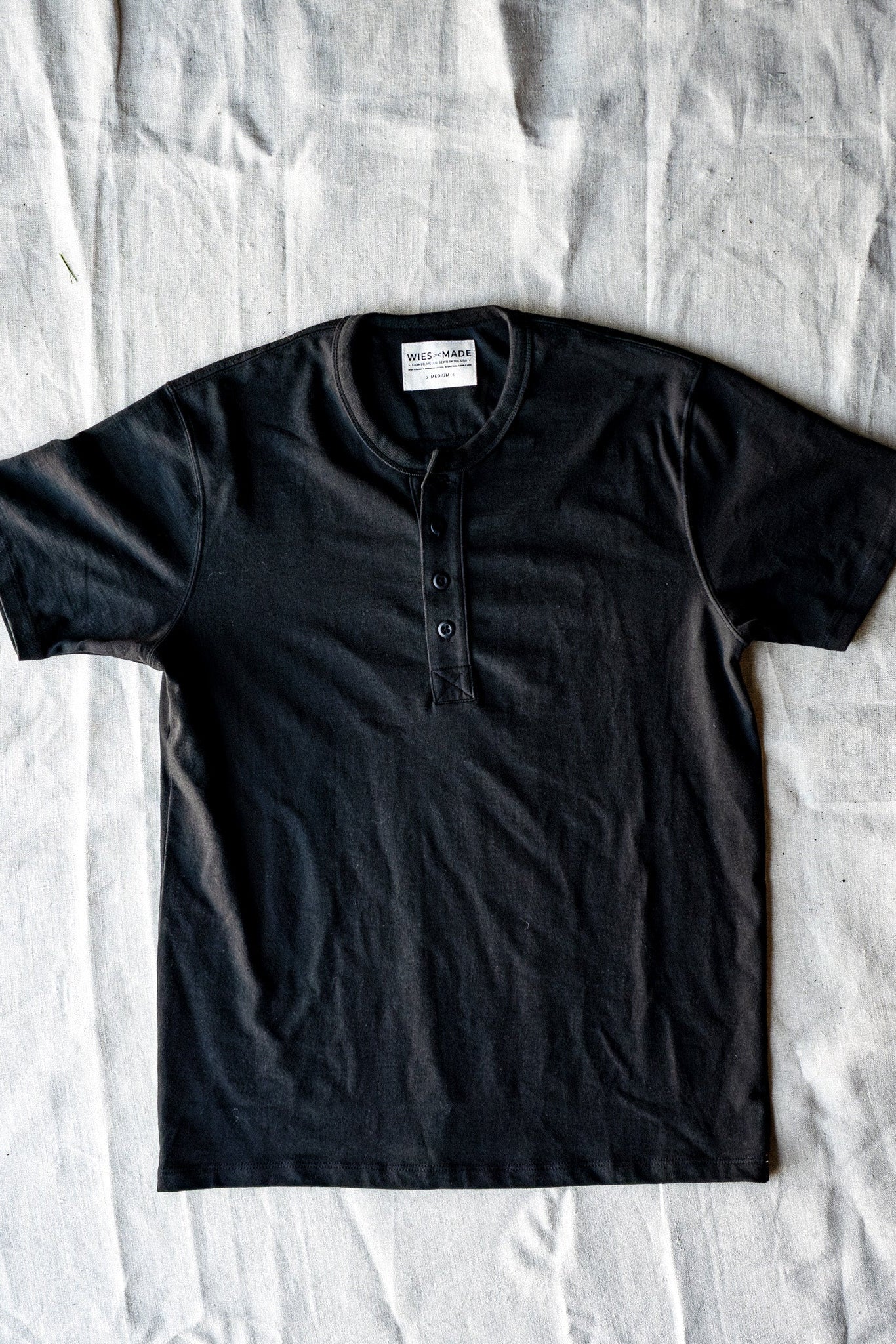Basalt black short-sleeve four-button Henley laid flat on natural linen, showing the WiesMade neck label.