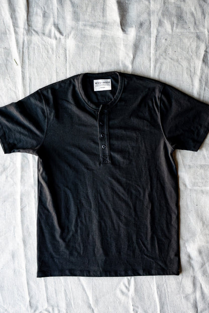Basalt black short-sleeve four-button Henley laid flat on natural linen, showing the WiesMade neck label.