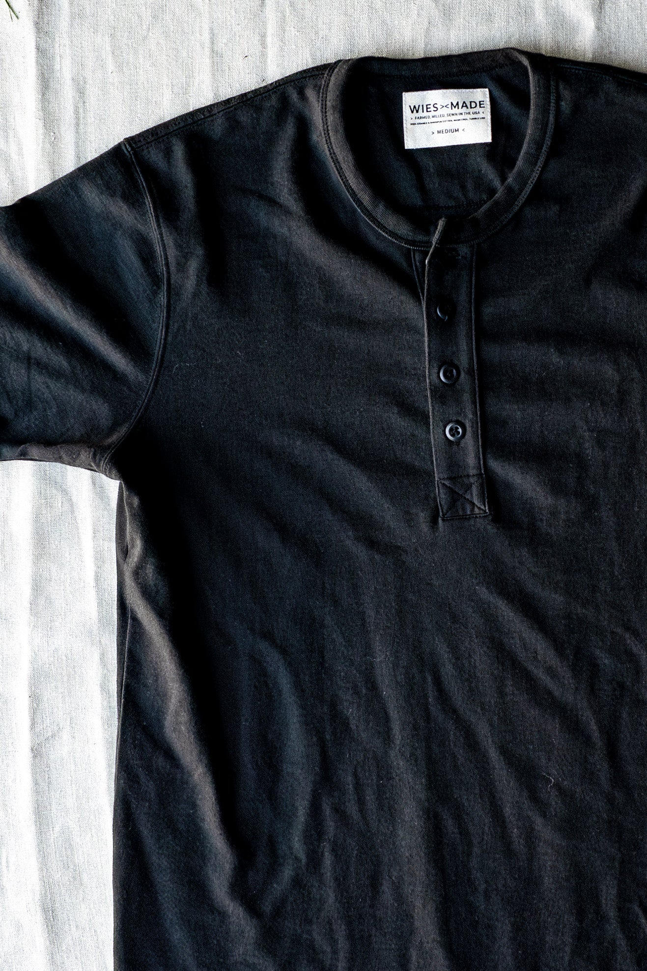 Basalt black four-button Bolinas Henley in heavyweight cotton, laid flat on linen with WiesMade neck label visible.