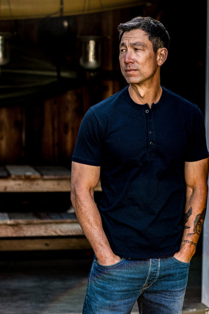 Man wearing a navy short-sleeve Bolinas Henley with four-button placket, paired with blue denim jeans in a rustic workshop.
