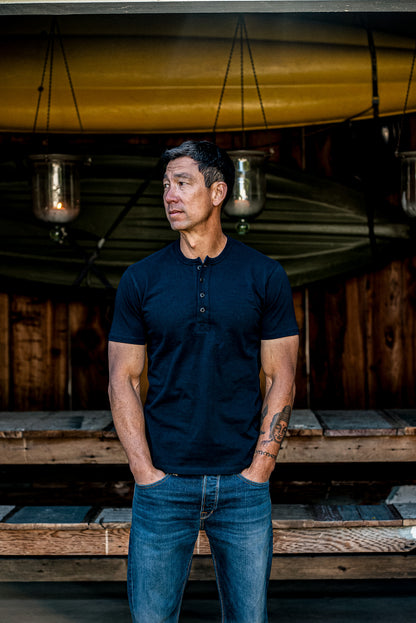 Man in a navy short-sleeve Bolinas Henley and blue jeans standing under a canoe on a wooden porch.