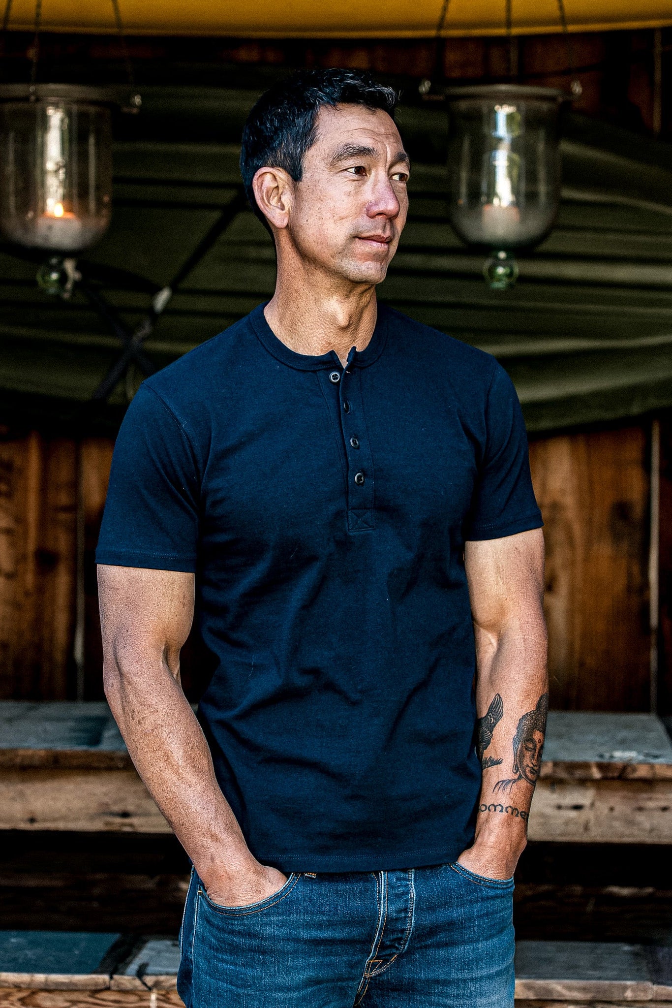 Model wears navy Bolinas short-sleeve Henley with denim jeans by a rustic wood cabin.