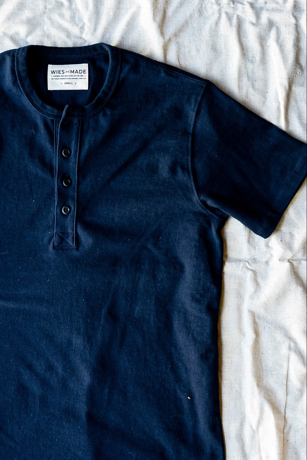 Navy short-sleeve four-button henley in heavyweight cotton laid flat on cream linen, WiesMade neck label visible.