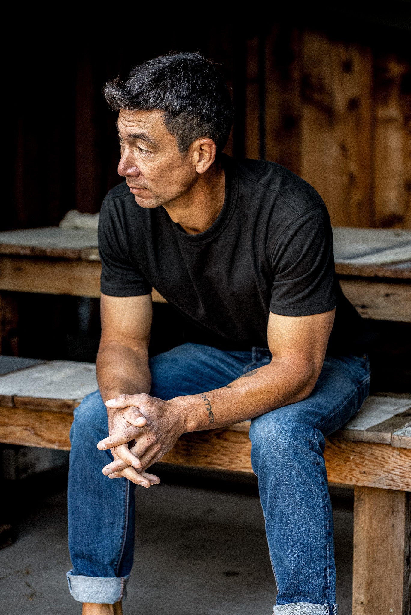 Man in a basalt black Bolinas heavyweight tee and cuffed blue jeans seated on a wooden workbench.
