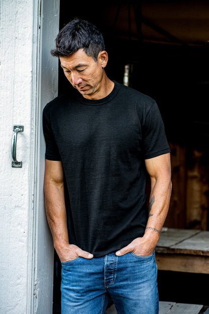 Man in a black Bolinas heavyweight tee and blue jeans leaning against a weathered white doorway.