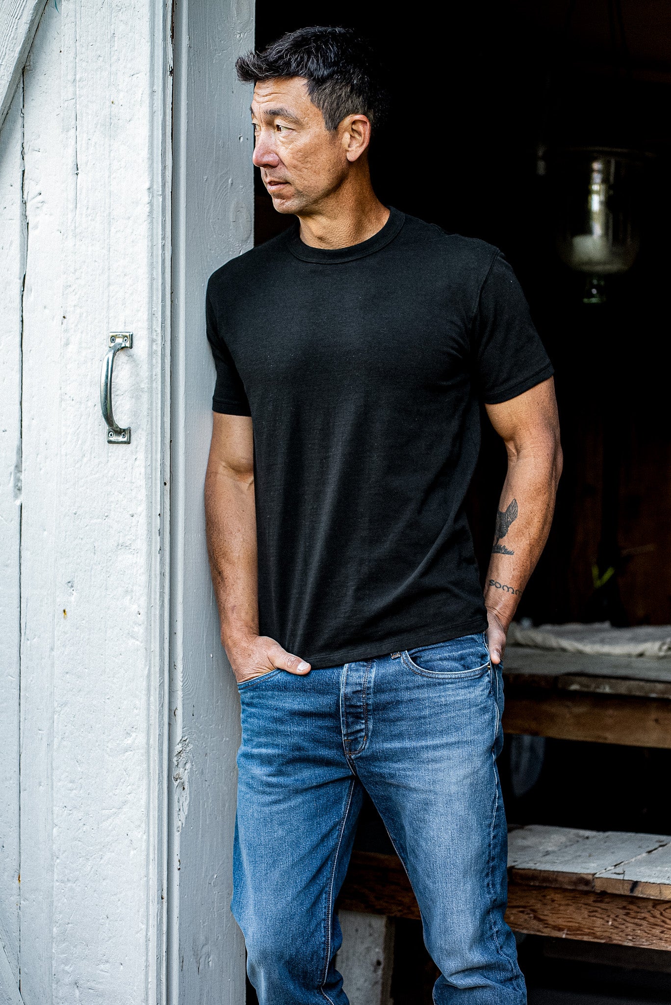 Man in a black heavyweight Bolinas tee and blue jeans leaning against a weathered white doorway.
