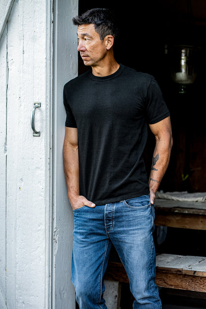 Man in a black heavyweight Bolinas tee and blue jeans leaning against a weathered white doorway.