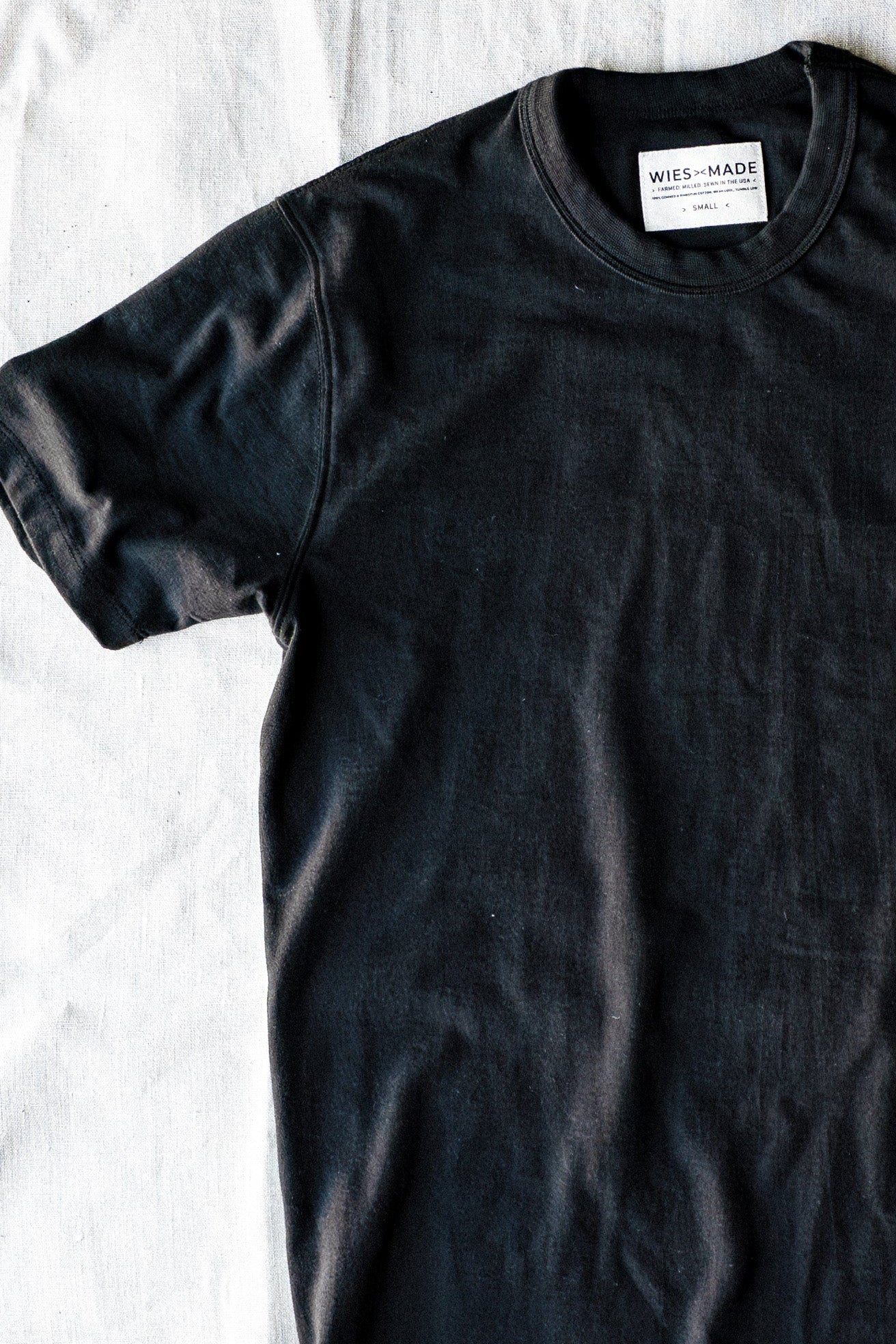 Basalt black heavyweight cotton Bolinas tee laid flat on linen, showing crewneck and WiesMade label.
