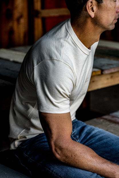 Side view of a man in a natural cotton Bolinas heavyweight tee and blue denim jeans, seated in a wooden workshop.