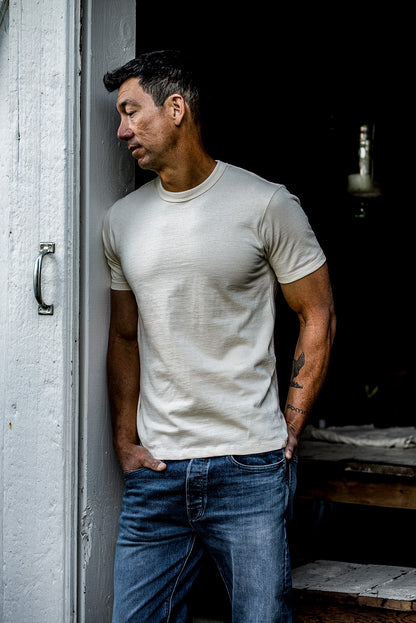 Man in a natural cotton Bolinas heavyweight tee and blue jeans leaning against a white doorframe.