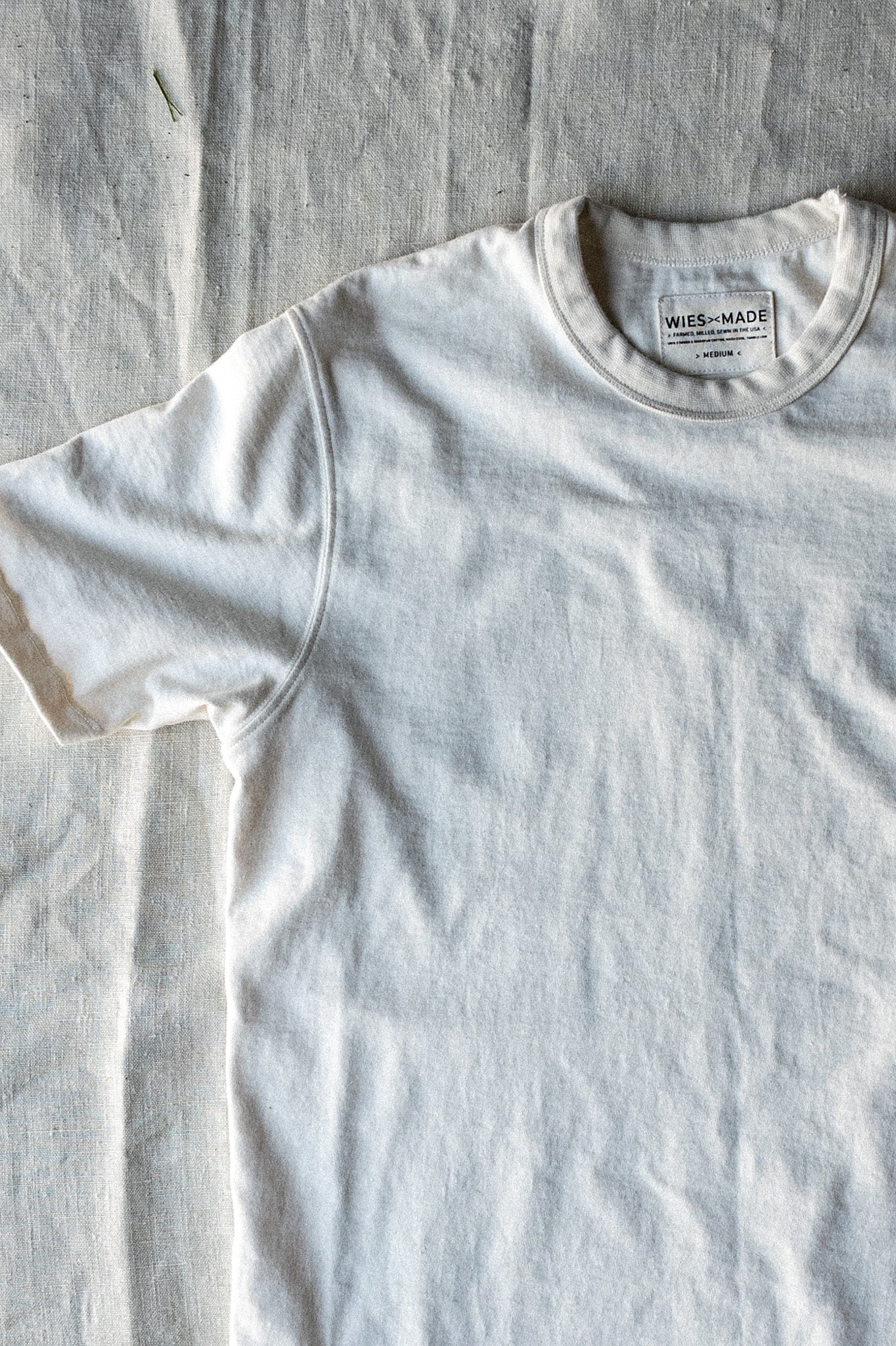 Natural cotton Bolinas heavyweight tee laid flat on linen, showing crewneck, shoulder seam, and WiesMade label.