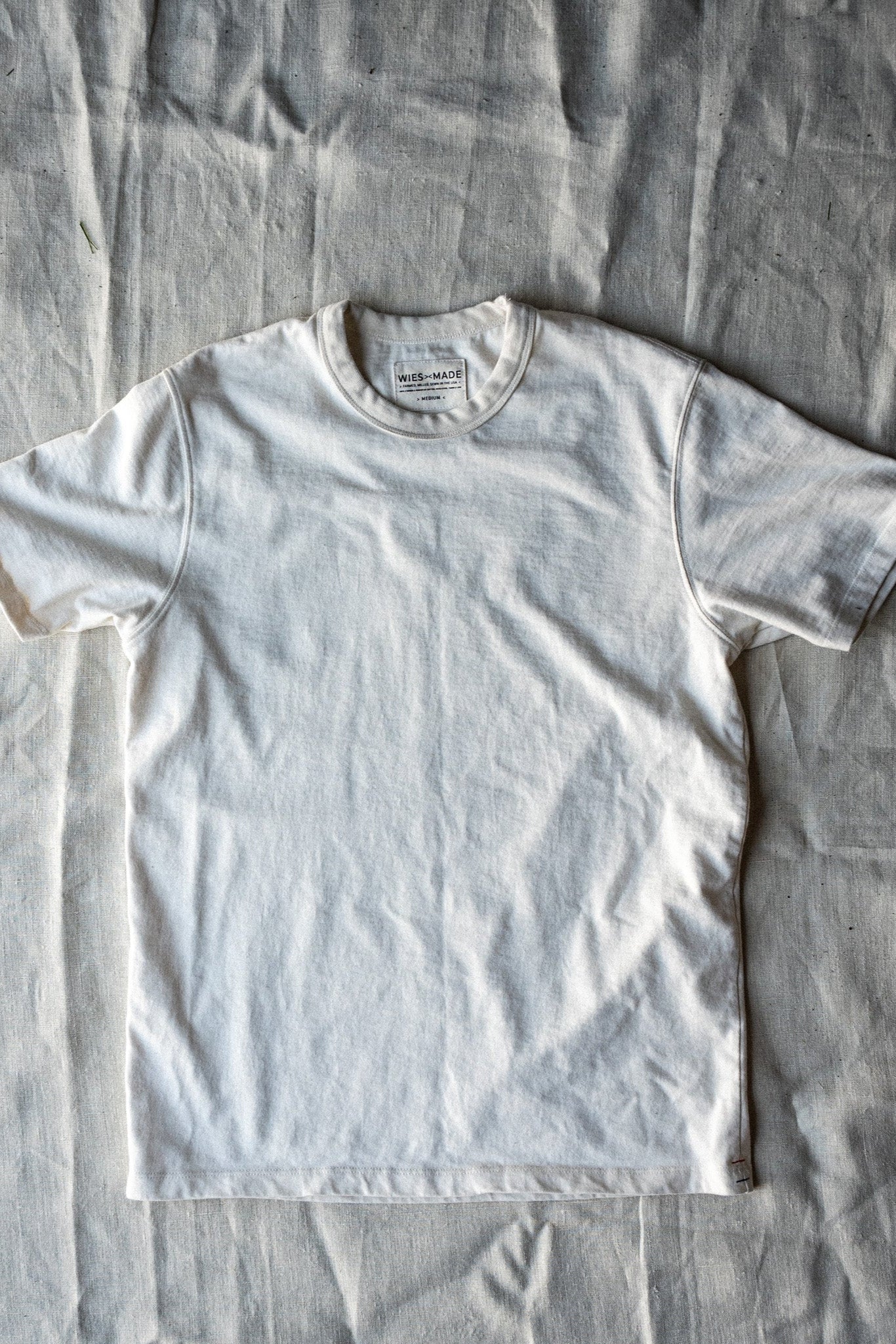 Natural white heavyweight cotton crewneck tee laid flat on a linen cloth, showing the WiesMade neck label.